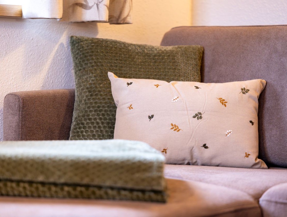 Ein beiger Kissen mit Blattmuster liegt auf einem braunen Sofa neben einem grünen Kissen