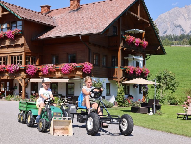 Kinder fahren Gokarts vor einem mit Blumen geschmückten Holzhaus in den Bergen