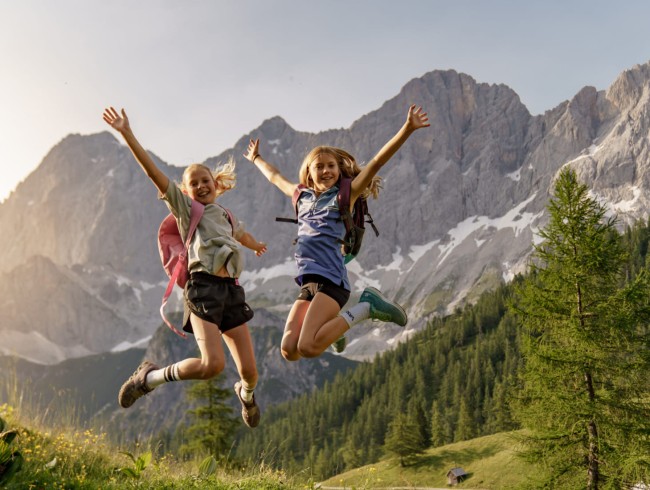 Zwei Mädchen springen fröhlich vor einer beeindruckenden Bergkulisse in die Luft