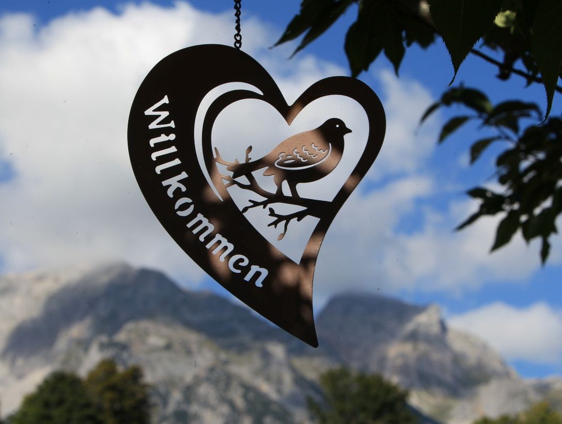 Herzf&ouml;rmiges Schild mit Vogelmotiv und "Willkommen", im Hintergrund Berge und Himmel